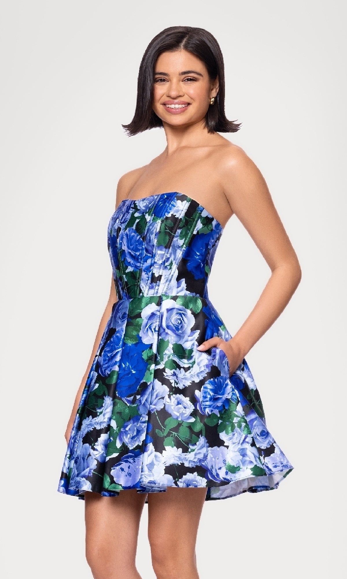 A woman smiles in the Strapless Print Hoco Dress: Blondie Nites 5592BN, a budget-friendly semi-formal strapless party dress featuring a blue and green floral pattern on white and ending at the knee.
